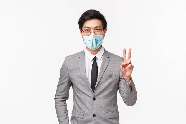 Porträtt av framgångsrik beslutsam och säker ung asiatisk man känsla han kan få affären för företag, står i grå kostym, visar fred tecken och leende, stående vit bakgrund — Stockfoto