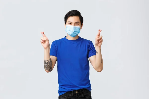 Olika känslor, social distans, självkarantän på covid-19 och livsstil koncept. Beslutsam hoppfull asiatisk man be, kors fingrar lycka, bär medicinsk mask — Stockfoto