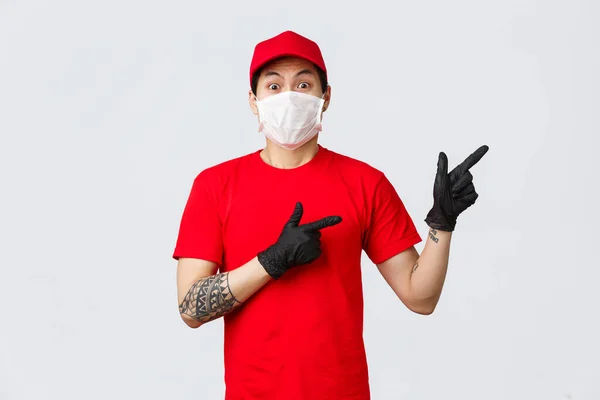 Šokovaný a vyděšený asijský poslíček v lékařské masce, ochranných rukavicích a červené uniformě ukazující prstem doprava. Obezřetný tai kurýr v červeném tričku a čepici, z čehosi nervózní, ukazující cestu — Stock fotografie