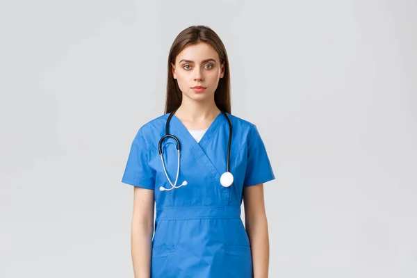 Pracownicy służby zdrowia, zapobiegać wirusom, ubezpieczenia i koncepcji medycyny. poważnie wyglądająca młoda pielęgniarka, lekarz w niebieskim fartuchu i stetoskopie, patrzący w kamerę, stojące szare tło — Zdjęcie stockowe