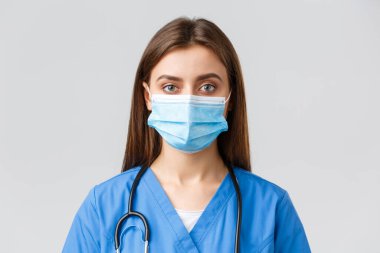 Covid-19, virüsün önlenmesi, sağlık, sağlık çalışanları ve karantina konsepti. Yakın plan genç bayan hemşire ya da doktor mavi önlüklü ve tıbbi maskeli kameraya ciddi bakıyor.
