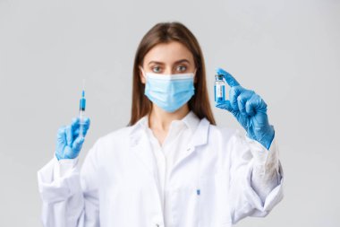 Covid-19, virüsü önleme, sağlık çalışanları ve karantina konsepti. Tıbbi maske takan doktor, lastik eldiven ve ameliyat önlüğü, ampulü koronavirüs aşısıyla gösteriyor, şırıngayı tut, hastaya iğne yap.