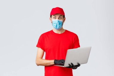Müşteri desteği, covid-19 teslimat paketleri, online sipariş işleme konsepti. Şaşırmış kurye kırmızı üniformalı, maske ve eldiven takıyor, kameraya bakıyor, dizüstü bilgisayar tutuyor, ilginç haberler okuyor.