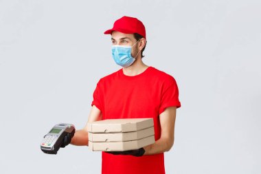 Yemek teslimatı, başvuru, çevrimiçi bakkal, temassız alışveriş ve covid-19 konsepti. Kırmızı üniformalı, eldivenli ve maskeli hoş teslimatçı. Posta terminali müşterisine pizza dağıtıyor.