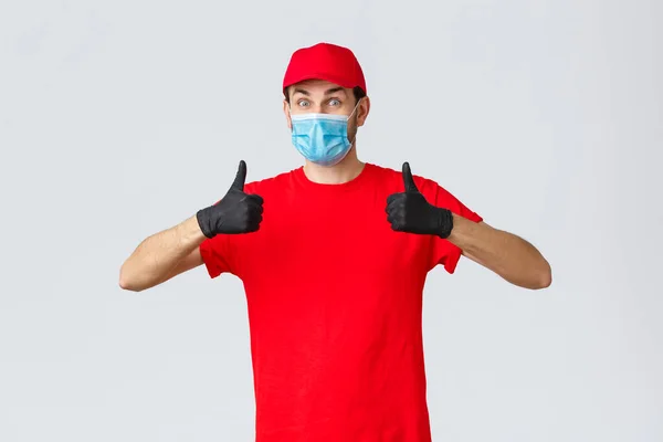 Covid-19, concetto di auto-quarantena, shopping e spedizione. Corriere entusiasta e felice in uniforme rossa, guanti e maschera viso, idea di supporto, thumb-up, consiglia la consegna del pacchetto senza contatto con il cliente — Foto Stock