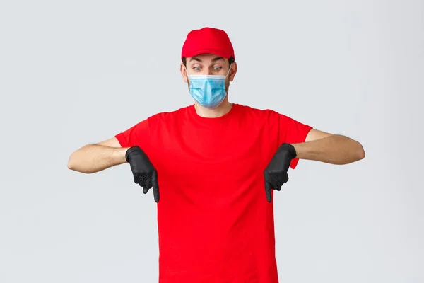 Covid-19, auto-quarantaine, achats en ligne et concept d'expédition. Courrier surprenant et choqué en uniforme, bonnet rouge et t-shirt, portant des mesures de sécurité masque médical et gants, pointant vers le bas — Photo