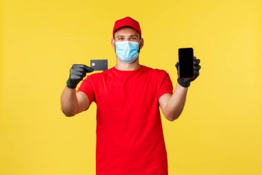 Yemek teslimatı, izleme emirleri, covid-19 ve kendini karantina konsepti. Tıbbi maske ve eldiven takan gülümseyen kurye, kredi kartı ve akıllı telefon gösteriyor, market alışverişini ödemenin yollarını gösteriyor.