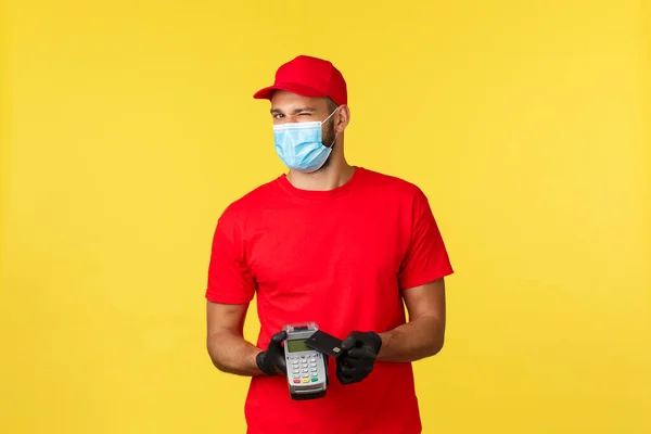 Food delivery, tracking, covid-19 and self-quarantine concept. Friendly courier in red uniform, medical mask and gloves, wink at camera, asking pay contactless with payment terminal and credit card