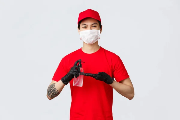 Souriant livreur en bonnet rouge et t-shirt, porter un masque médical et des gants, appliquer un désinfectant pour les mains à l'abri de la covide-19, virus pendant l'épidémie. Courrier se soucie des clients, utiliser un désinfectant pendant le travail — Photo