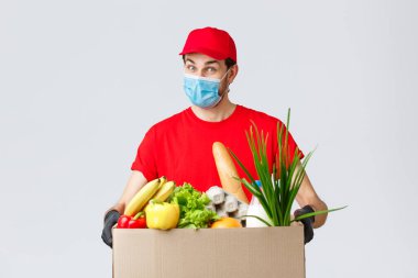 Erzak ve paket teslimatı, covid-19, karantina ve alışveriş konsepti. Yüz maskeli ve eldivenli dost canlısı kurye, kırmızı üniformalı. İnternetten sipariş edilen müşteriye yiyecek kutusu getir.