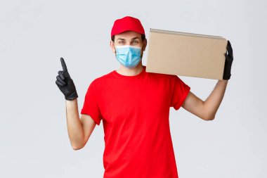 Paketler ve paket teslimatı, karantina teslimatı, nakil emirleri. Yüz maskeli neşeli kurye, eldivenler ve üniforma, paketini kapının önüne taşıyın, yukarı doğru, sırayla kutuyu tutun.