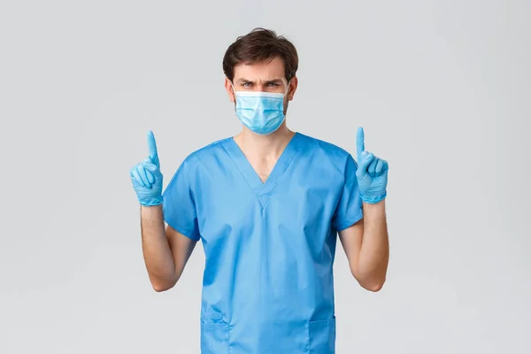 Covid-19, egészségügyi dolgozók és kórházi koncepció. Komoly férfi orvos kék köpenyben, orvosi maszk és kesztyű, ujjal felfelé mutogatva, fontos jelzést vagy híreket mutatva, top hirdetés — Stock Fotó