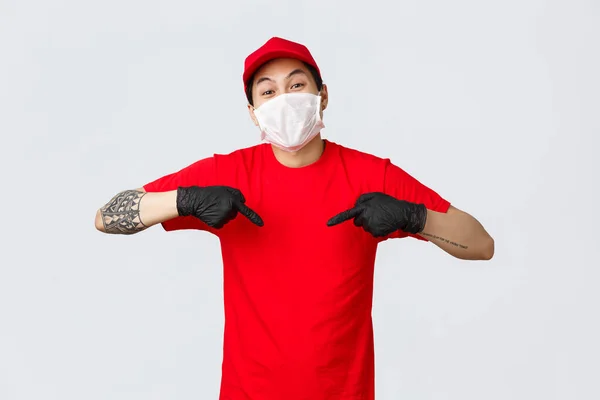 I tid leverans med detta företag kurirtjänst. Glada asiatiska kurir i mössa och röd t-shirt, bär medicinsk mask och handskar för att skydda kunderna från covid19 virus spridning, pekar sig — Stockfoto