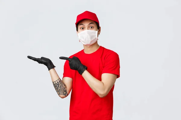 Regarde-moi ça. Homme de livraison asiatique excité en bonnet rouge, t-shirt, portant des gants de protection en latex et un masque médical, montrant quelque chose en main, pointant vers la paume et la caméra de regard impressionné — Photo