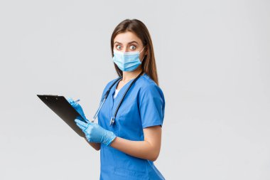 Covid-19, virüsün önlenmesi, sağlık, sağlık çalışanları ve karantina konsepti. Şok olmuş ve ürkmüş kadın hemşire ya da doktor mavi önlüklü, tıbbi maskeli, elinde pano bakışlarıyla kameraya şaşırmış.