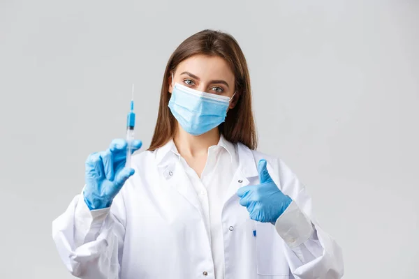 Covid-19, 예방 바이러스, 의료 종사자 및 격리 개념. 의료용 마스크와 고무장갑을 착용 한 결단력있는 젊은 의사는 코로나 바이러스 백신으로 엄지와 주사기를 보여 주고 있다. — 스톡 사진