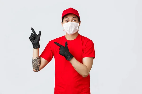 Surpreso e impressionado homem de entrega, asiático mensageiro em vermelho uniforme t-shirt e cap, máscara médica protetora e luvas para a segurança do cliente. Courier apontando dedos canto superior esquerdo no anúncio — Fotografia de Stock