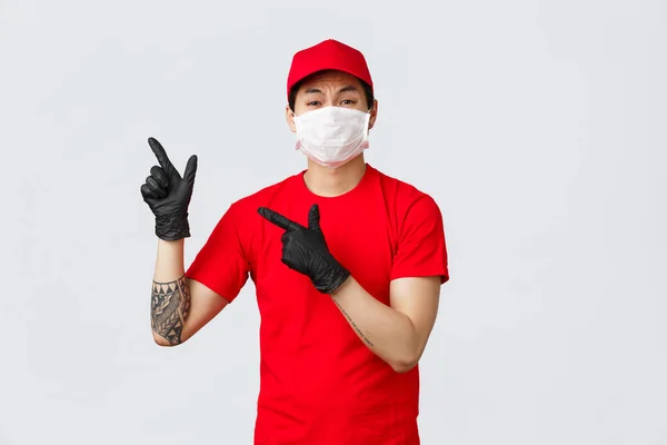 Bouleversé, déçu asiatique courrier se plaindre de la publicité. Livraison homme en t-shirt rouge et casquette, portant un masque médical et des gants de protection pendant la covid19, les doigts pointant vers le coin supérieur gauche — Photo