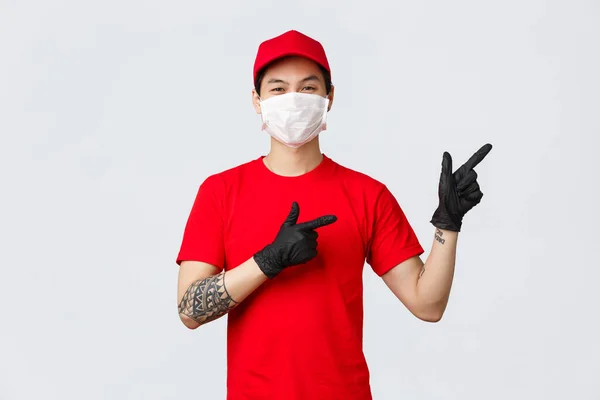 Klikněte na toto tlačítko, stáhněte si doručovací aplikaci. Asijský kurýr v červené uniformě čepice a tričko, nosí ochrannou masku, lékařské rukavice, ukazující prsty vpravo, pozvat vyzkoušet promo, představit speciální nabídku — Stock fotografie