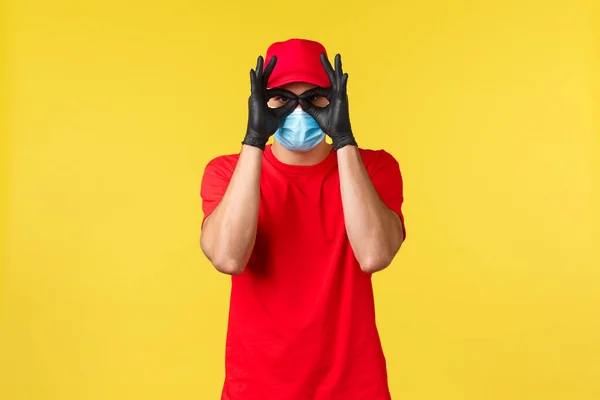 Экспресс-доставка во время пандемии, covid-19, безопасная доставка, концепция онлайн-покупок. Смешной курьер в красной униформе, медицинской маске и перчатках, показывает окаи или очки с руками — стоковое фото