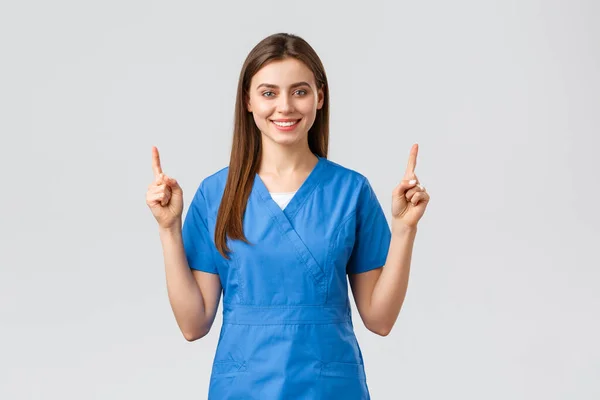 Healthcare workers, prevent virus, insurance and medicine concept. Attractive nurse or doctor in blue scrubs, pointing fingers up and smiling, advertise banner, medical pills, grey background — Stock Photo, Image