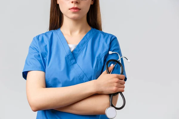 Healthcare workers, medicine, insurance and covid-19 pandemic concept. Cropped shot of young female doctor or nurse in blue scrubs with serious face, cross arms and hold stethoscope — Stock Photo, Image
