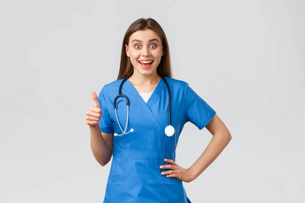 Trabalhadores médicos, cuidados de saúde, covid-19 e conceito de vacinação. Surpreendido excitado enfermeira ou médico em azul esfrega com estetoscópio, mostrar polegar-se em aprovação, como ideia, sorrindo otimista — Fotografia de Stock