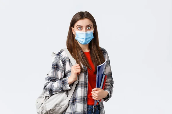 Coronavirus pandemic, covid-19 education, and back to school concept. Shocked and surprised girl in medical mask, student gasping over big news in campus, hold notebooks and backpack