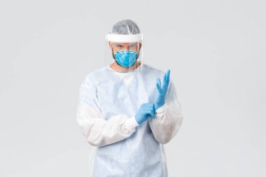 Covid-19 salgını, virüs salgını, klinik ve sağlık çalışanları konsepti. Ciddi görünümlü koruyucu ekipmanlı kararlı bir doktor, lastik eldiven takın ve kendinden emin görünün.