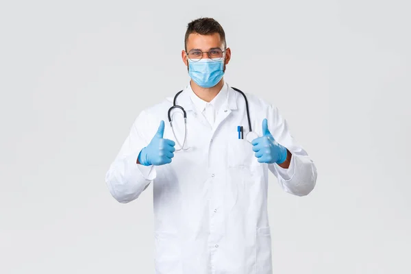 Covid-19, zdravotničtí pracovníci, pandemie a prevence viru. Optimistický usmívající se lékař v bílých pláštích a lékařské masce, ukázat palce nahoru v souhlasu, podpůrný lékařský program — Stock fotografie