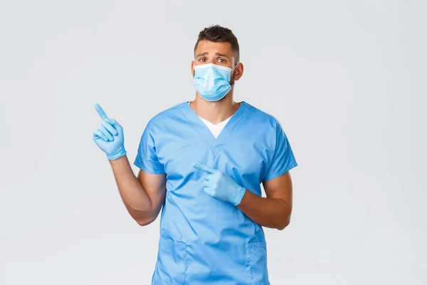 Gezondheidswerkers, covid-19, coronavirus en het voorkomen van virus concept. Onhandige trieste knappe dokter met medisch masker en scrubs informeren slecht nieuws, wijzende vingers linksboven en kijken boos — Stockfoto