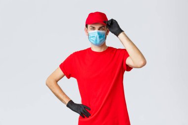 Covid-19, kendini karantina, online alışveriş ve nakliye konsepti. Teslimatçı kırmızı üniformalı, şapkalı müşteriyi selamlıyor, koronavirüs salgınında çalışıyor, tıbbi maske takıyor ve koruyucu eldiven takıyor.
