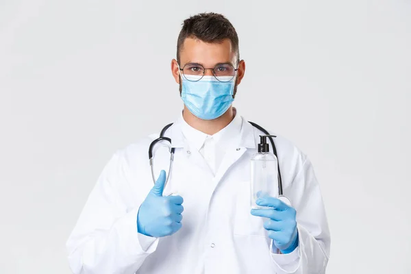 Covid-19, healthcare workers, pandemic and preventing virus concept. Close-up of confident serious doctor in medical mask and gloves, thumb-up advice good hand sanitizer, protect from coronavirus