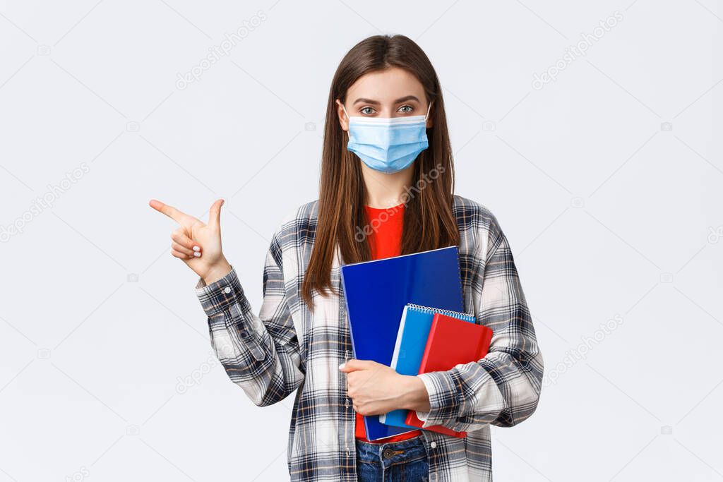 Coronavirus pandemic, covid-19 education, and back to school concept. Young pretty female student in medical mask with notebooks, pointing finger left, showing university info