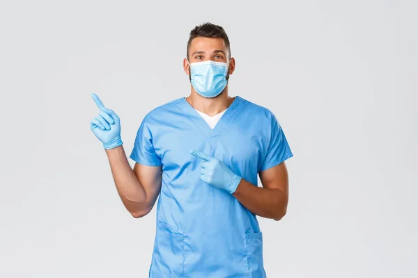 Hälso- och sjukvårdspersonal, covid-19, coronavirus och förebyggande av virus koncept. Glad professionell manlig sjuksköterska, läkare i skrubb och medicinsk mask, handskar pekar finger vänster, visar banner — Stockfoto