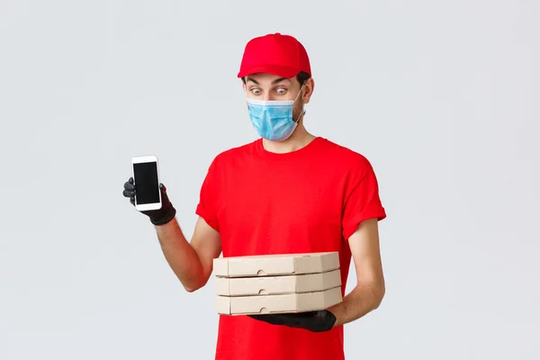 Consegna di cibo, applicazione, generi alimentari online, shopping senza contatto e concetto di covid-19. Divertente corriere divertito in uniforme rossa, maschera viso e guanti, mostrando app schermo smartphone e tenere scatole di pizza — Foto Stock