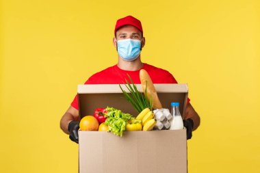 Paket teslimat, COVid-19 karantina ve market konsepti. Kırmızı üniformalı ve tıbbi maskeli mutlu kurye, evden alışveriş yapmak için müşteri evine market kutusunu getiriyor.
