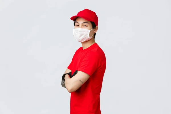 Pewny siebie azjatycki dostawca w czerwonej czapce, maska medyczna i rękawiczki, profil stojący z ramionami skrzyżowanymi klatki piersiowej, obrócić się w kamerze z pewnym siebie wyglądem, zapewnić bezpieczną i szybką pracę przewoźnika, koncepcja kuriera — Zdjęcie stockowe