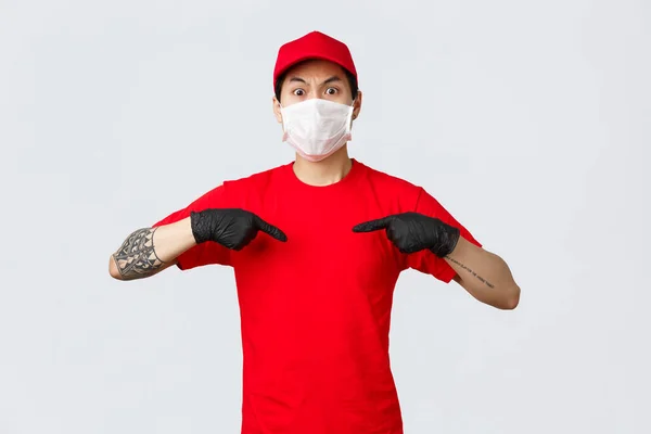 Scioccato e interrogato uomo consegna asiatica in uniforme berretto rosso e t-shirt, indossando maschera medica protettiva e guanti. Fornire servizio in situazione di pandemia di quarantena, puntare se stesso gonfio — Foto Stock