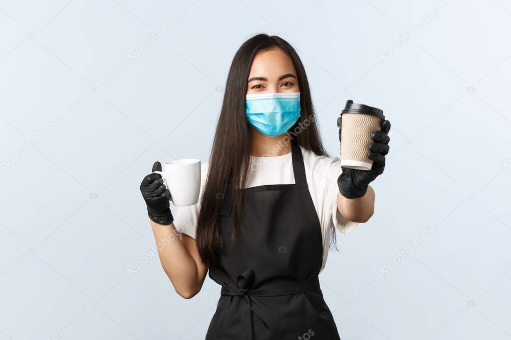Covid-19, social distancing, small coffee shop business and preventing virus concept. Cute smiling asian barista recommend using takeaway paper cups during coronavirus, handing you drink