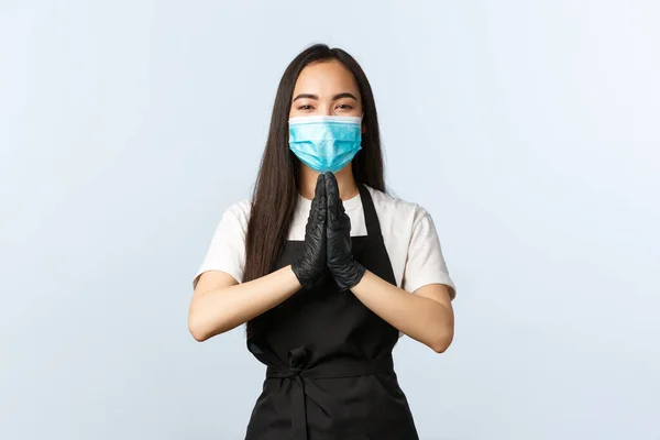 Covid-19, social distancing, small coffee shop business and preventing virus concept. Cute smiling asian employee, cafe barista in medical mask and gloves, hold hands in pleading gesture, pray