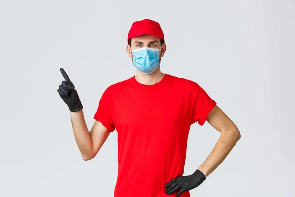 Covid-19, vlastní karanténa, on-line nakupování a expedice koncept. Pošťák v červené čepici a tričku, lékařská maska s rukavicemi na ochranu klientů a zaměstnanců, ukazováček vlevo na promo, ukaž reklamu — Stock fotografie