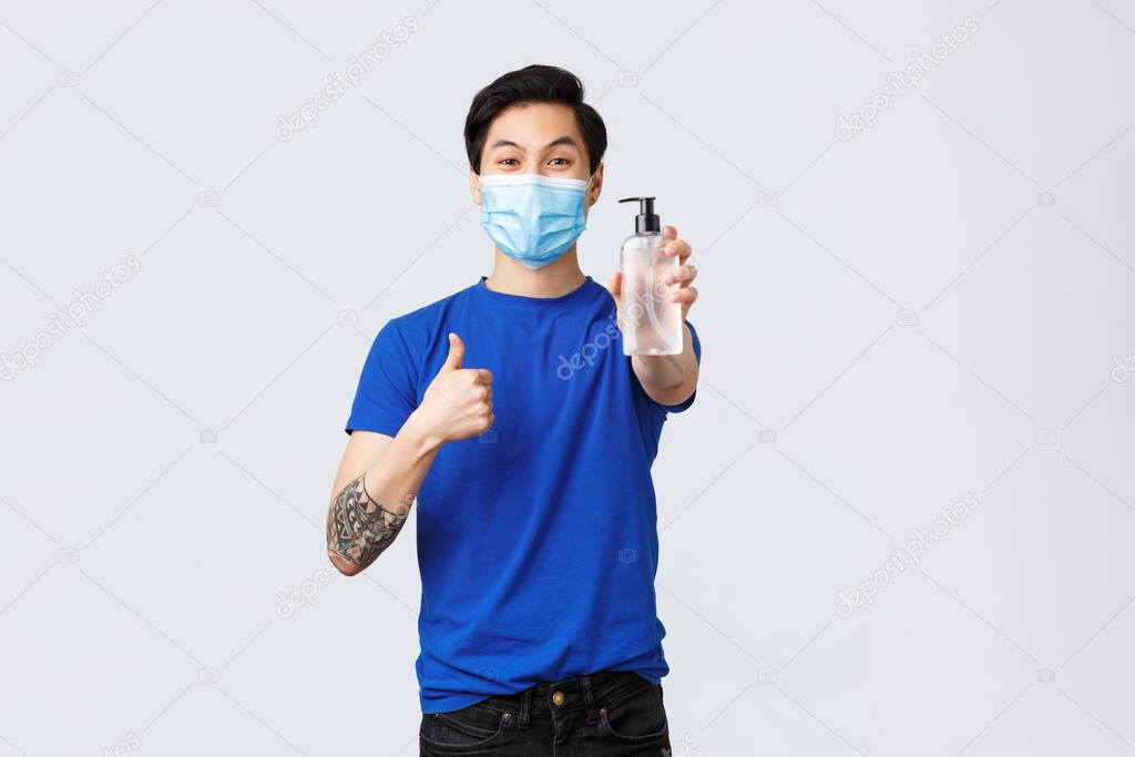 Lifestyle, people different emotions and covid-19 pandemic concept. Friendly good-looking asian guy in medical mask explain importance using hand sanitizer during coronavirus, show thumb-up
