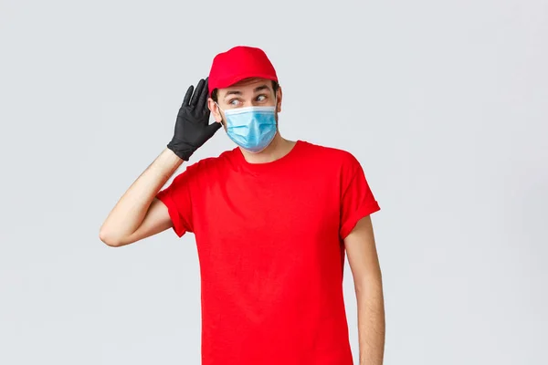 Groceries and packages delivery, covid-19, quarantine and shopping concept. Intrigued courier in red uniform, face mask and gloves, overhearing conversation, eavesdrop, hold hand near ear — Stock Photo, Image