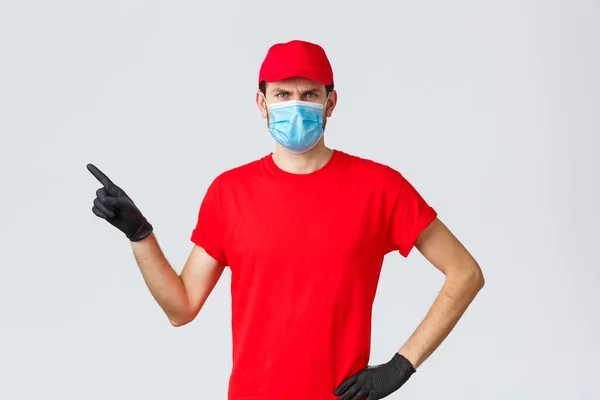 Covid-19, self-quarantine, online shopping and shipping concept. Displeased angry delivery guy scolding courier making mistake in transfer, frowning, pointing finger left, wear medical mask — Stock Photo, Image