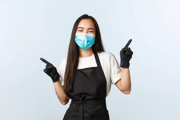 Covid-19, social distancing, small coffee shop business and preventing virus concept. Friendly smiling, happy asian cafe employee in medical mask and gloves pointing sideways, show two charts