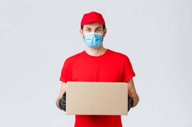 Paketler ve paket teslimatı, covid-19 kendi kendini karantinaya alma, transfer emirleri. Kırmızı üniformalı genç kurye, eldivenler ve yüz maskesi, tutucu kutusu, müşteriye dağıtım emri, irtibatsız servis.