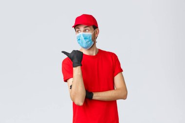 Covid-19, kendini karantina, online alışveriş ve nakliye konsepti. Kırmızı üniformalı, yüz maskeli, eldivenli, parmakla işaret eden ve soldan etkilenmiş, ilginç bir şekilde tanıtım kitabı okuyan,