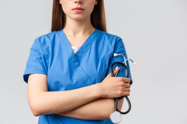 Sağlık çalışanları, ilaç, sigorta ve covid-19 salgını konsepti. Ciddi yüzlü, çapraz kollu ve steteskoplu mavi önlüklü genç bayan doktor ya da hemşire.