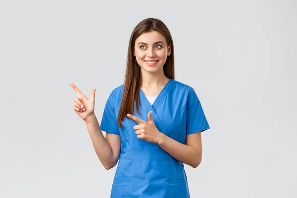Pracownicy służby zdrowia, zapobiegać wirusom, ubezpieczenia i koncepcji medycyny. Uśmiechnięta ładna pielęgniarka lub lekarz w niebieskim fartuchu, wskazując palcami w lewo i patrząc na baner z uśmiechem, szary tło — Zdjęcie stockowe